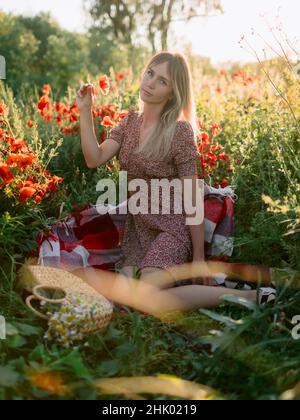 Donna bionda all'aperto in plaid con fiori di papavero al tramonto. Donna in abito e borsa con fiori da campo Foto Stock