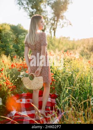 Donna bionda all'aperto in plaid con fiori di papavero al tramonto. Donna in abito e borsa con fiori da campo Foto Stock