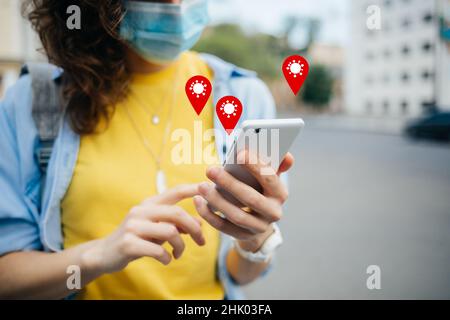 Concetto dell'applicazione di monitoraggio COVID-19 con icone di geolocalizzazione a comparsa. Donna che indossa maschera protettiva che tiene smartphone con software di tracciamento coronavirus o Foto Stock