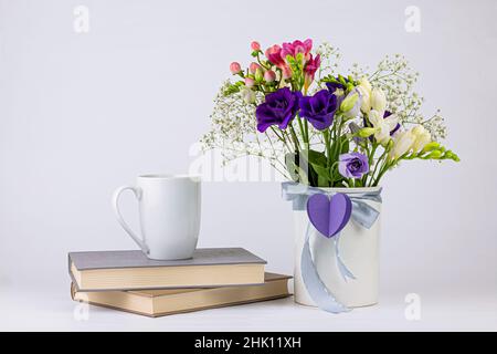 Colori diversi bouquet di fiori belli in vaso bianco d'epoca con nastro e cuore di carta, tazza di caffè o tè e libri su sfondo bianco. Morn Foto Stock
