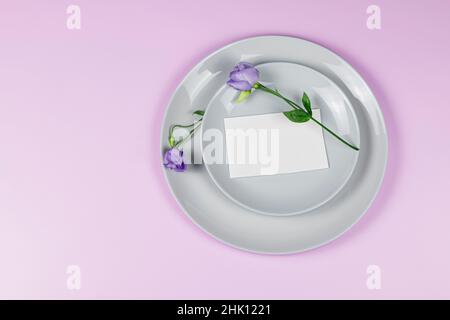 Festa saluto mockup scena. Biglietti d'auguri vuoti, piatti grigi, e fiori viola rami di eustoma su sfondo rosa con spazio copia. Disposizione piatta, a. Foto Stock