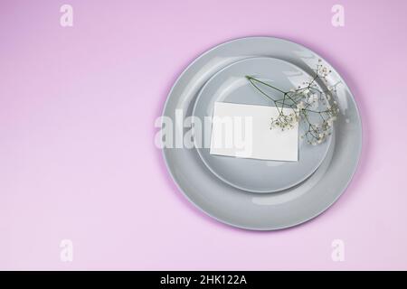 Festa saluto mockup scena. Biglietti d'auguri vuoti, piatti grigi, e fiori bianchi rami gypsophila su sfondo rosa con spazio copia. Disposizione piatta, Foto Stock