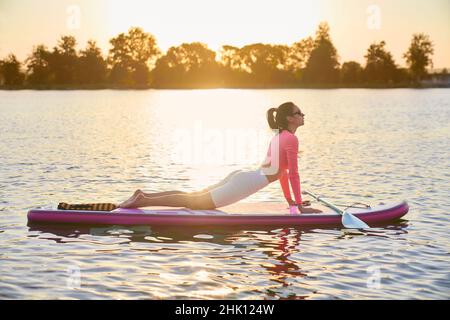 Bella donna attiva che fa esercizi di fitness su tavola sup con incredibile tramonto estivo sullo sfondo. Sport acquatici e concetto di persone. Foto Stock