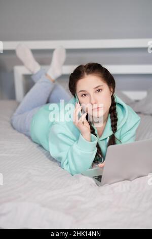 Formazione a distanza online e lavoro. Felice brunette donna ragazza lavoro ufficio lavoro a distanza da casa in turchese felpa con cappuccio e jeans a letto. Telefono in conversazione. Utilizzando un computer portatile Foto Stock