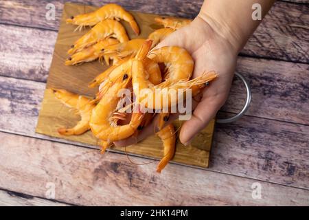 la mano di una donna tiene una manciata di gamberi bolliti sullo sfondo di un tavolo di legno Foto Stock