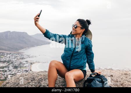 Vista laterale dell'escursionista femminile che prende un selfie e sorride mentre si siede su una roccia Foto Stock
