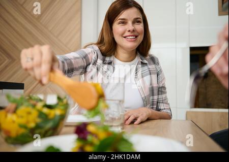 Bella donna caucasica attraente, casalinga, servendo una sana e deliziosa insalata vegana cruda su un piatto bianco e sorrisi seduti a tavola in casa sua Foto Stock