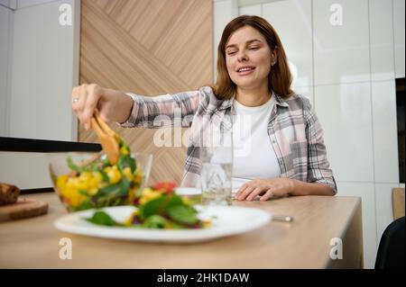 Bella donna caucasica, casalinga, servendo una sana e deliziosa insalata vegana cruda su un piatto bianco e sorrisi seduti a tavola in casa sua Foto Stock