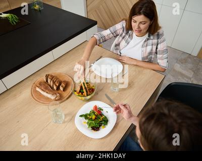 Direttamente sopra, bella giovane donna incinta, moglie attenta che serve suo marito un'insalata di verdure. Felice coppia caucasica godere di insieme oltre l Foto Stock