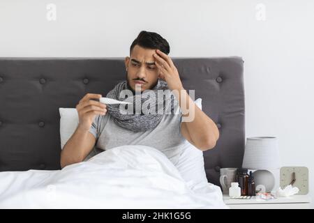Uomo mediorientale malato indossando sciarpa seduta in letto e termometro di tenuta Foto Stock