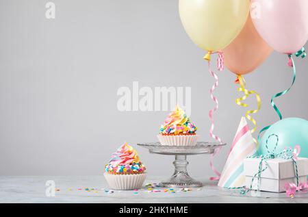 Due cupcake di compleanno dell'arcobaleno con regali, cappelli e palloncini colorati su sfondo grigio chiaro. Scena da una festa di compleanno! Foto Stock