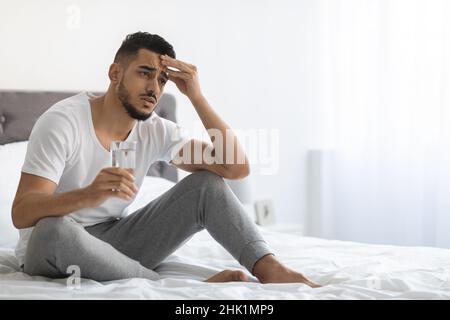 Concetto di hangover. Sick Arab Guy che tiene un bicchiere d'acqua e tocca la testa Foto Stock