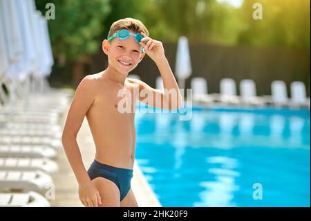 Ragazzo che tocca gli occhiali che sorridono alla macchina fotografica vicino alla piscina Foto Stock