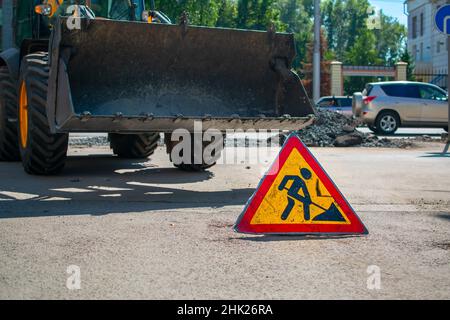 il simbolo dei lavori su strada è installato sullo sfondo di un trattore, un apripista con benna sollevata Foto Stock