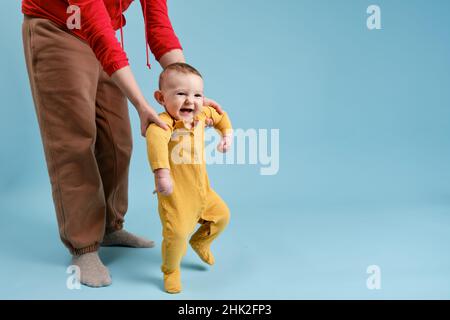 La madre insegna al bambino a stare in piedi sui piedi e a camminare, il bambino su sfondo blu studio Foto Stock