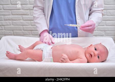 Il medico controlla la temperatura del neonato con un termometro. Un infermiere in uniforme misura la febbre del bambino con un termometro Foto Stock