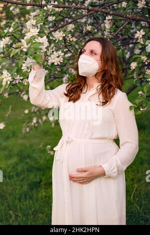 Donna incinta in maschera medica n95 con fiori di melo in primavera natura. Felicità della gravidanza durante l'epidemia di covid, pandemia di coronavirus q Foto Stock