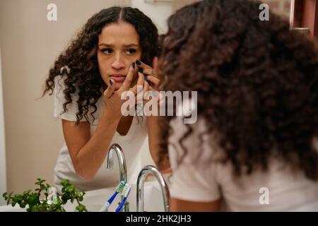 La donna dell'America Africana spreme i difetti sul viso mentre si trova accanto allo specchio del bagno Foto Stock