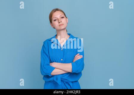 Ritratto fotografico giovane donna esperta con mani ripiegate, con camicia blu, con espressione contemplativa, in piedi accanto ad uno spazio vuoto, isolato su sfondo blu pastello. Foto Stock