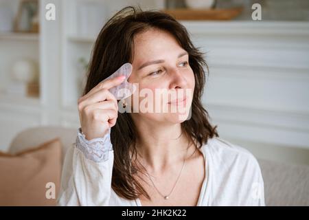 Cute donna caucasica con scraper gouache di giada in soggiorno sul divano in accappatoio, donna brunetta che fa un massaggio viso con un scraper gouache. Bellezza e cura personale, concetto di cura della pelle e del viso Foto Stock