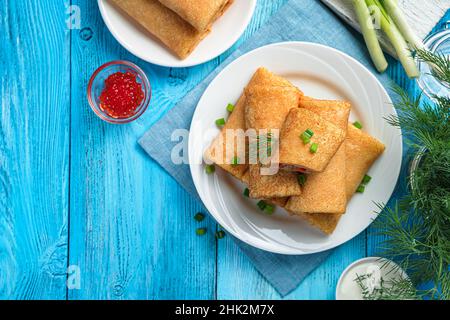 Pancake ripieni decorati con aneto e cipolla con caviale rosso, panna acida su sfondo blu. Frittelle tradizionali russe fritte. Vista dall'alto, copia sp Foto Stock