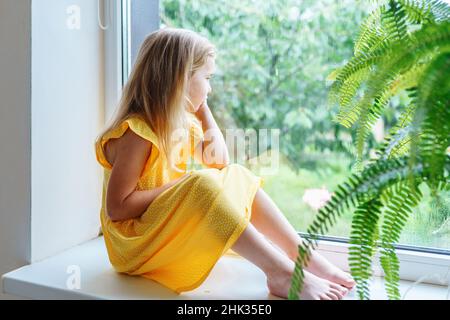 Triste annoiato bambina sul davanzale che guarda fuori la finestra nel giorno d'estate piovoso Foto Stock