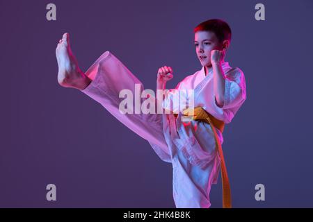 Ritratto di ragazzo sportivo, taekwondo maschio, karate atleti in doboks che fanno movimenti di base isolato su sfondo viola in neon. Concetto di sport Foto Stock