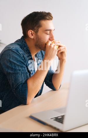 Colpo verticale di uomo libero affamato mangiare hamburger guardare video online sul computer portatile durante la pausa. Foto Stock