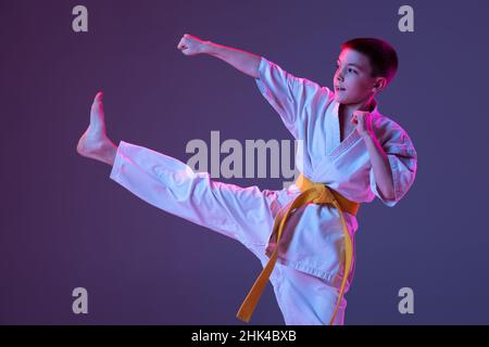 Ritratto di ragazzo sportivo, taekwondo maschio, karate atleti in doboks che fanno movimenti di base isolato su sfondo viola in neon. Concetto di sport Foto Stock