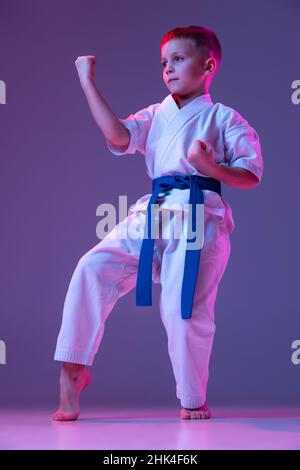 Ritratto di ragazzo sportivo, taekwondo maschio, karate atleti in doboks che fanno movimenti di base isolato su sfondo viola in neon. Concetto di sport Foto Stock