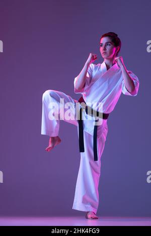 Ritratto di taekwondo femminile, karate atleti in doboks che fanno movimenti di base isolato su sfondo viola in neon. Concetto di sport, arti marziali Foto Stock