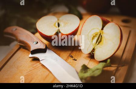 Su un tagliere di legno si trova una deliziosa mela rossa matura, tagliata a due metà, e accanto ad essa si trova un coltello affilato. Nutrizione sana. Dieta. Gallina di frutta Foto Stock