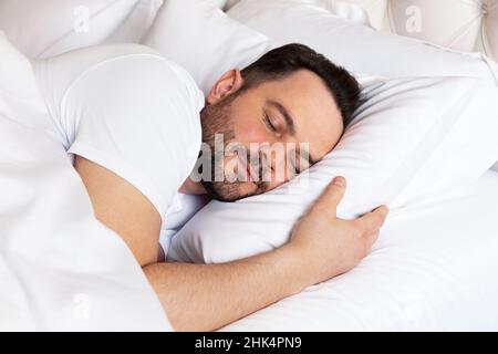 Bel giovane uomo che dorme su biancheria da letto bianca. Vista di un bell'uomo sorridente che dormiva nel letto di casa. Foto Stock