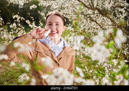 Donna allergica che soffre di allergia stagionale in primavera, sorridendo in giardino fiorito in primavera, utilizzando gocce nasali tra alberi in fiore. Concetto di allergia primaverile Foto Stock