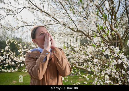 Donna allergica che soffre di allergia stagionale in primavera, posando in giardino fiorito in primavera. Giovane donna starnutita di fronte a un albero in fiore. Concetto di allergia primaverile Foto Stock