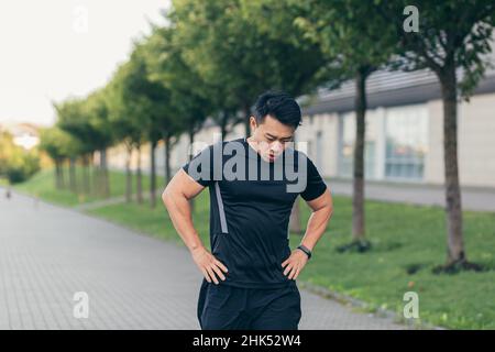 L'atleta asiatico maschile, stanco dopo uno jogging mattutino, corre nel parco vicino allo stadio Foto Stock