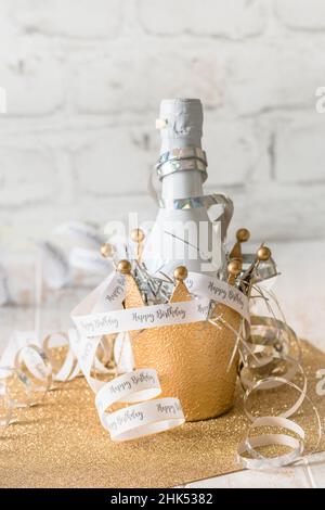 Piccola bottiglia bianca con vino frizzante in una corona decorativa in metallo dorato, decorata da streamers con testo Happy Birthday, verticale con spazio copia Foto Stock