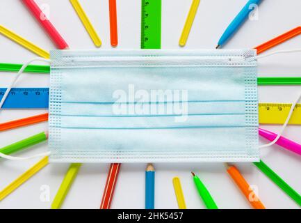 Sfondo della scuola con maschera medica su forniture multicolore: Penne, matite, righelli. Vista dall'alto. Concetto di scuola o quarantena. Spazio di copia per il testo. Foto Stock