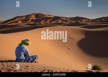 Un uomo berbero in abito tradizionale siede tra le dune di sabbia di Erg Chebbi, deserto del Sahara, Marocco, Nord Africa, Africa Foto Stock