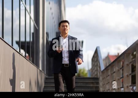 Il capo asiatico dell'uomo d'affari guarda il telefono mobile legge le notizie dal telefono e scrive nel messaggero fuori dell'ufficio Foto Stock