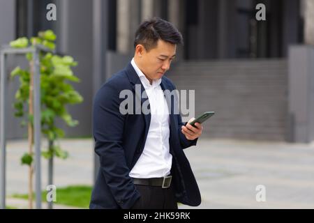 Il capo asiatico dell'uomo d'affari guarda il telefono mobile legge le notizie dal telefono e scrive nel messaggero fuori dell'ufficio Foto Stock