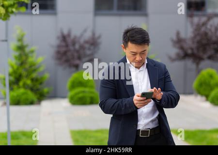 Il capo asiatico dell'uomo d'affari guarda il telefono mobile legge le notizie dal telefono e scrive nel messaggero fuori dell'ufficio Foto Stock