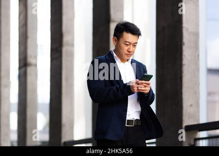 Il capo asiatico dell'uomo d'affari guarda il telefono mobile legge le notizie dal telefono e scrive nel messaggero fuori dell'ufficio Foto Stock