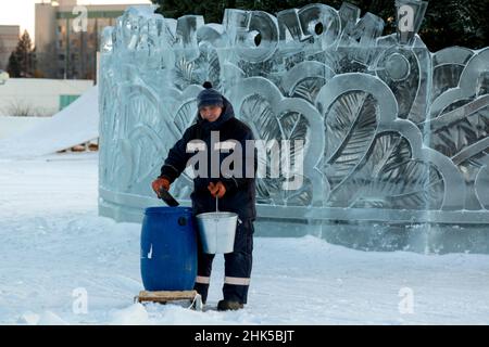 Un lavoratore con un secchio in mano attira l'acqua da un barile blu con una mestolo nel luogo di montaggio della città di ghiaccio Foto Stock