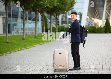 Felice turista asiatico con una grande valigia vicino all'aeroporto, ad un incontro d'affari arriva guardando la macchina fotografica e sorridendo Foto Stock
