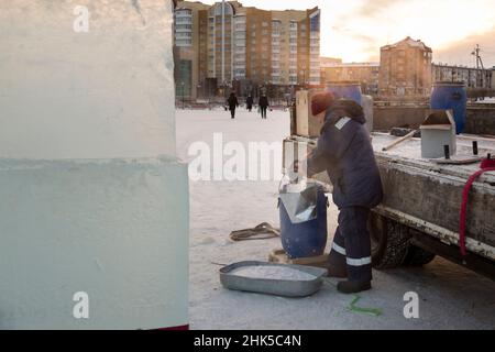 Un lavoratore con un secchio in mano attira l'acqua da un barile blu con una mestolo nel luogo di montaggio della città di ghiaccio Foto Stock