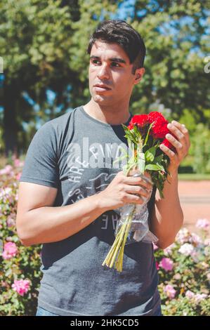 giovane latino caucasico ispanico fuori nel parco con un bouquet di fiori in mano, triste, pensante e confuso guardando lato, verticale. Foto Stock