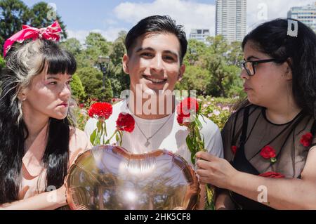 giovane latino caucasico uomo in un parco pubblico, seduto sorridendo nervoso con fiore e un cuore palloncino guardando la macchina fotografica e due donne arrabbiate guardando Foto Stock