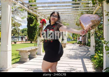 giovane ragazza latina caucasica ispanica godendo giorno di valentine in un parco, sta ballando e tenendo un pallone a forma di cuore e un fiore in mano, lei Foto Stock