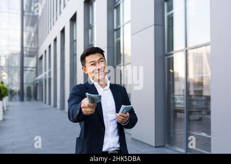 Uomo d'affari asiatico maschile gioisce tenendo un sacco di soldi guardando la fotocamera dando soldi Foto Stock
